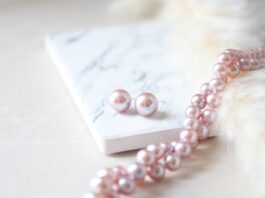 Pink pearl stud earrings beside a pink pearl necklace resting on a white marble-topped notebook.
